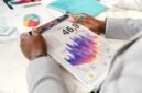 Seorang eksekutif bisnis sedang menganalisis grafik kinerja keuangan dalam laporan tahunan, yang menggambarkan upaya memahami arah dan strategi perusahaan. Gambar ini merepresentasikan pentingnya membaca data secara cermat untuk mengidentifikasi kisah di balik angka-angka perusahaan.