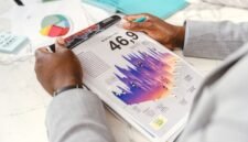 Seorang eksekutif bisnis sedang menganalisis grafik kinerja keuangan dalam laporan tahunan, yang menggambarkan upaya memahami arah dan strategi perusahaan. Gambar ini merepresentasikan pentingnya membaca data secara cermat untuk mengidentifikasi kisah di balik angka-angka perusahaan.