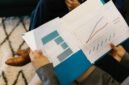 Beberapa profesional sedang menganalisis grafik dan data dari laporan tahunan perusahaan, mencerminkan proses mendalam dalam memahami kinerja dan merencanakan strategi bisnis. Interaksi tersebut menunjukkan pentingnya diskusi dan kolaborasi untuk menginterpretasi informasi kompleks yang disajikan dalam dokumen tersebut.