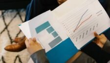 Beberapa profesional sedang menganalisis grafik dan data dari laporan tahunan perusahaan, mencerminkan proses mendalam dalam memahami kinerja dan merencanakan strategi bisnis. Interaksi tersebut menunjukkan pentingnya diskusi dan kolaborasi untuk menginterpretasi informasi kompleks yang disajikan dalam dokumen tersebut.