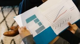 Beberapa profesional sedang menganalisis grafik dan data dari laporan tahunan perusahaan, mencerminkan proses mendalam dalam memahami kinerja dan merencanakan strategi bisnis. Interaksi tersebut menunjukkan pentingnya diskusi dan kolaborasi untuk menginterpretasi informasi kompleks yang disajikan dalam dokumen tersebut.