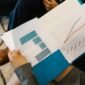 Beberapa profesional sedang menganalisis grafik dan data dari laporan tahunan perusahaan, mencerminkan proses mendalam dalam memahami kinerja dan merencanakan strategi bisnis. Interaksi tersebut menunjukkan pentingnya diskusi dan kolaborasi untuk menginterpretasi informasi kompleks yang disajikan dalam dokumen tersebut.